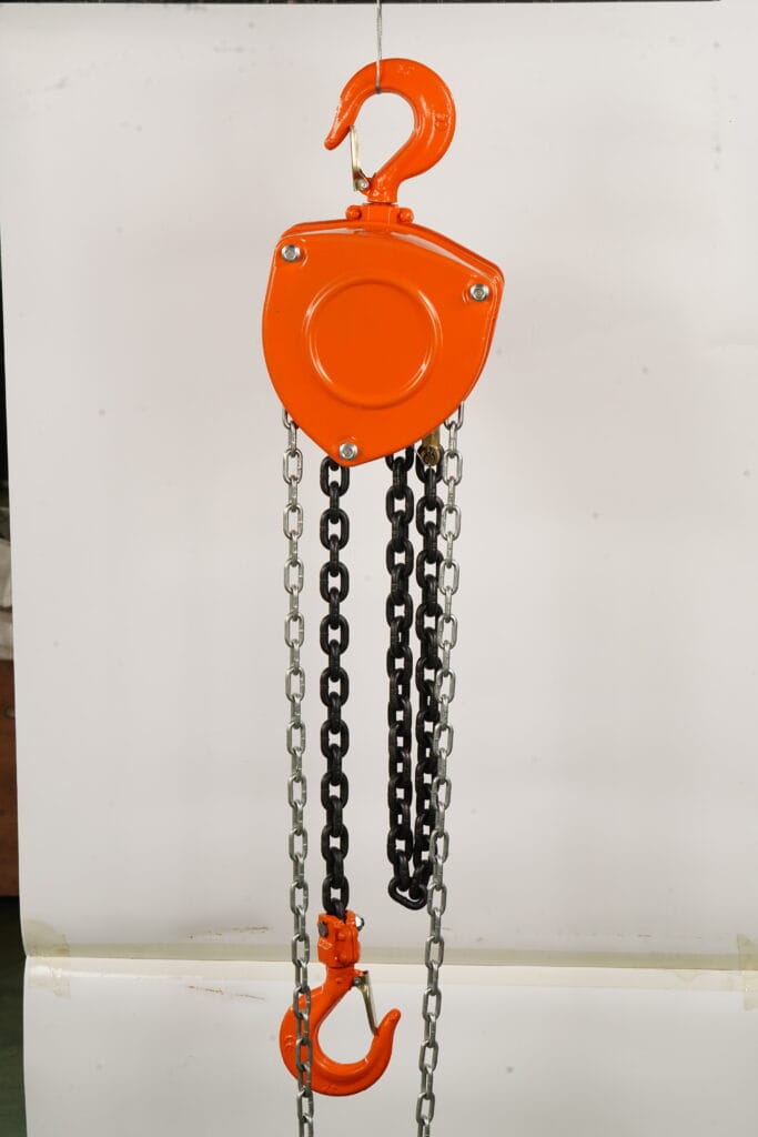 Bright orange triangular-body HSC manual chain hoist hanging from an overhead beam in a modern industrial workshop, with steel chains extended downward and clean metal structures in the background — showcasing light-duty lifting equipment for capacities from 0.5 to 20 tons.